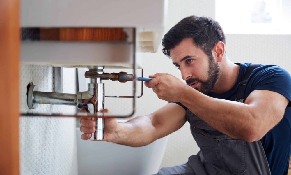 Male Plumber Using Wrench To Fix Leaking Sink In Home Bathroom